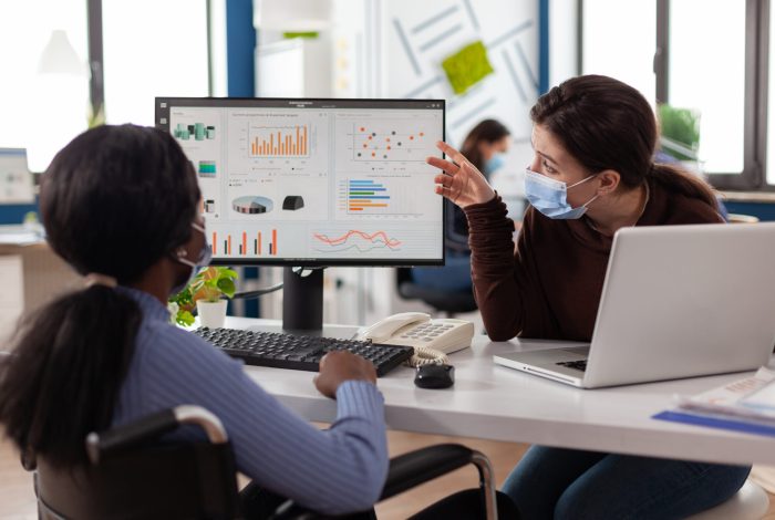 Multiethnic business team with medical face masks to prevent infection with coronavirus Multiethnic business team with medical face masks to prevent infection with coronavirus analyzing marketing statistics discussing strategy. Disabled paralyzed businesswoman working in startup office
